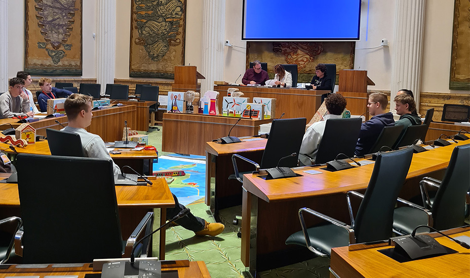 Leerlingen zitten op de stoelen (ronde setting) in de Statenzaal en luisteren naar de begeleider. In het midden worden met karton de onderwerpen uitgebeeld.